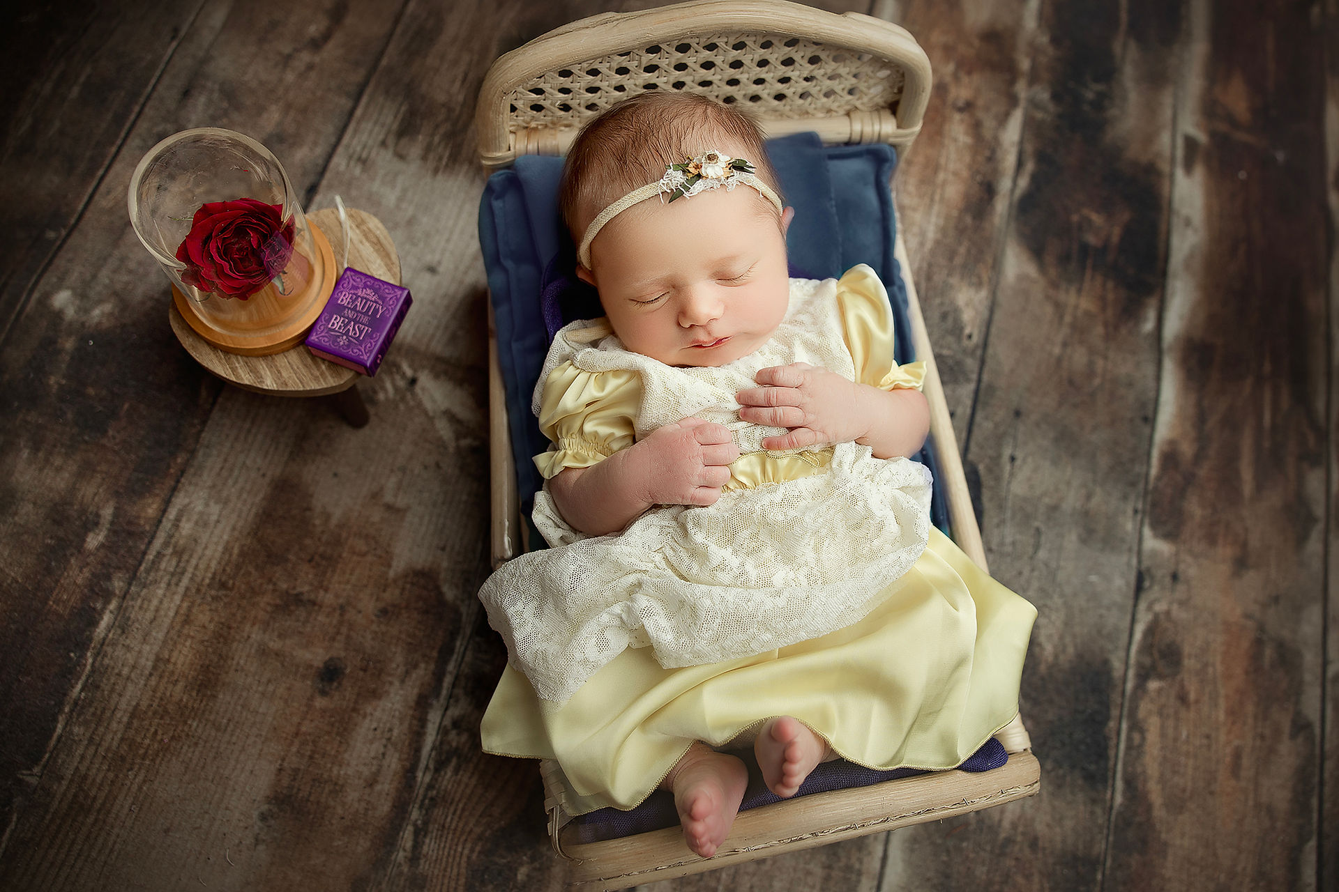 Newborn baby girl dressed as Belle from Beauty and the Beast, sleeping in a woven basket beside a tiny storybook and enchanted rose.
