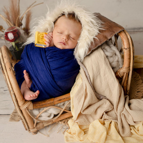 Newborn baby boy sleeping swaddled in deep blue fabric, styled as the Beast from Beauty and the Beast in a rustic basket setup.