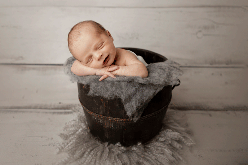 Aurora Colorado | Newborn Photographer | Tiny Details Photography | Newborn baby boy in luneberry water bucket with head on arms smiling with gray fur and landon white wood background