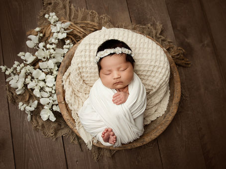 Top-down newborn portrait of a sleeping baby wrapped in a white swaddle, resting safely in a wooden bowl lined with soft cream fabric, surrounded by neutral textures and white florals.