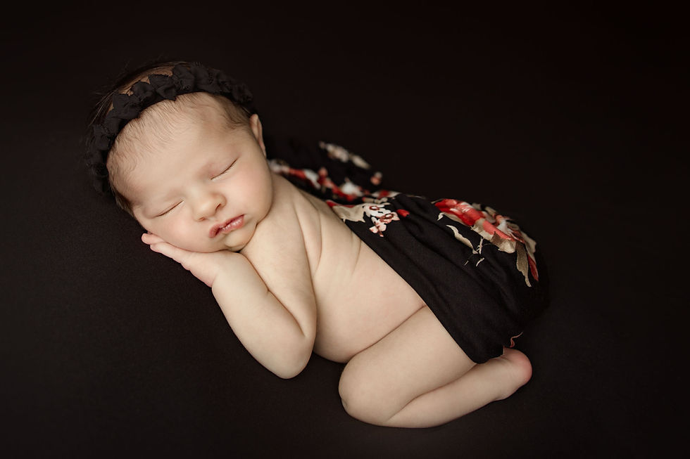 Sleeping newborn baby girl posed on her side against a dark backdrop, wearing a black floral wrap and soft headband.
