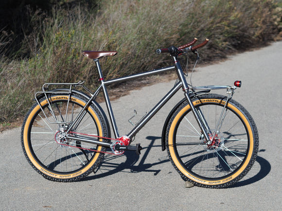 Titanium Nua Roure bike with a red Pinion P1.18 gearbox, Gates belt drive, 650x70mm tires, and custom stainless-steel racks.