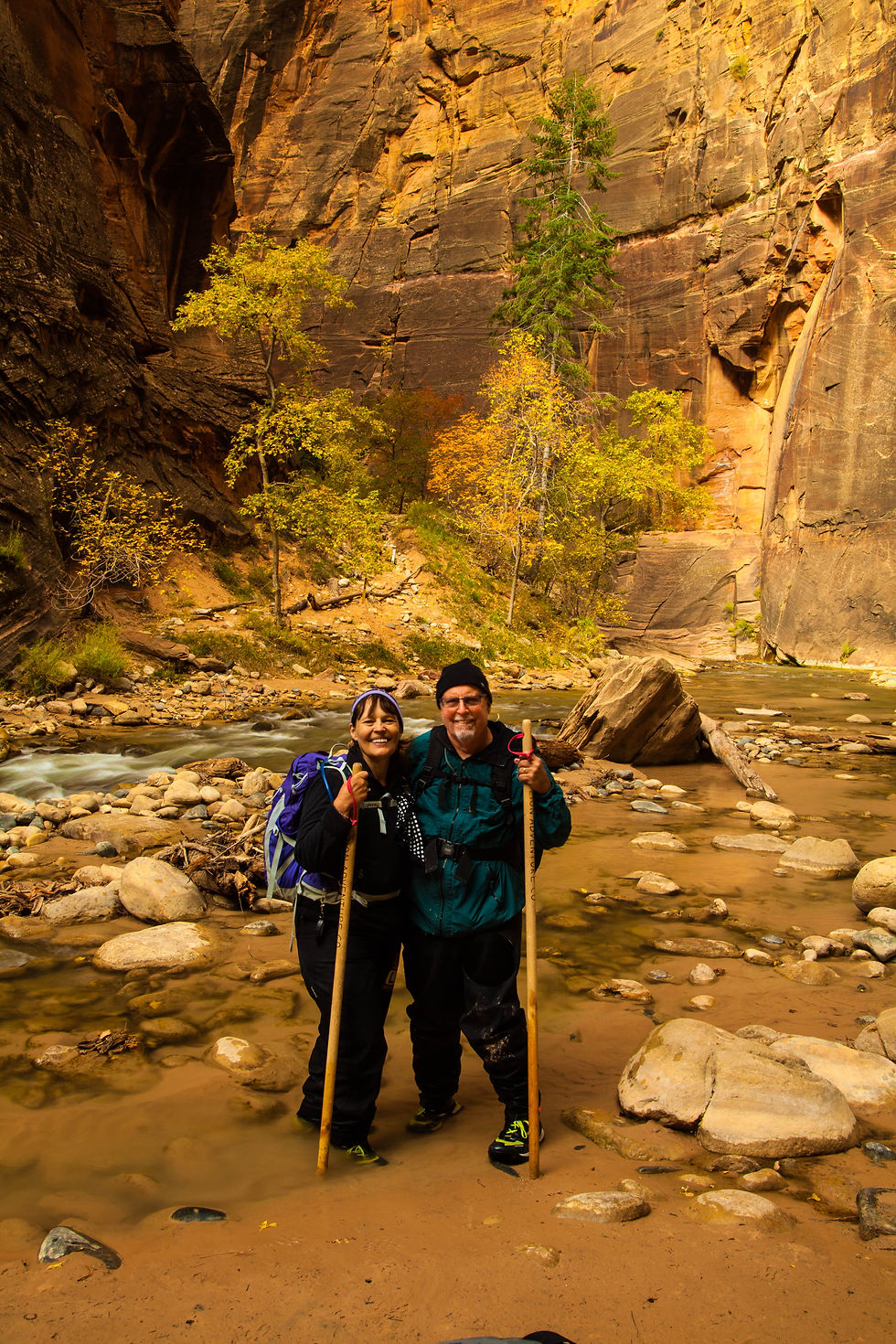 Lorna and Mick in the river with their hiking staffs and boulders in the river and red Cliffs in the background