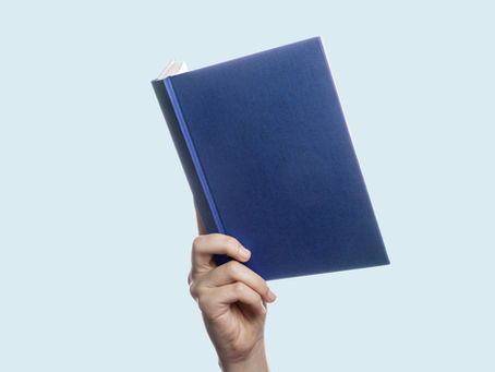 Hand holding up a blue hardcover book against a pale blue background, with a clean, minimalist feel.