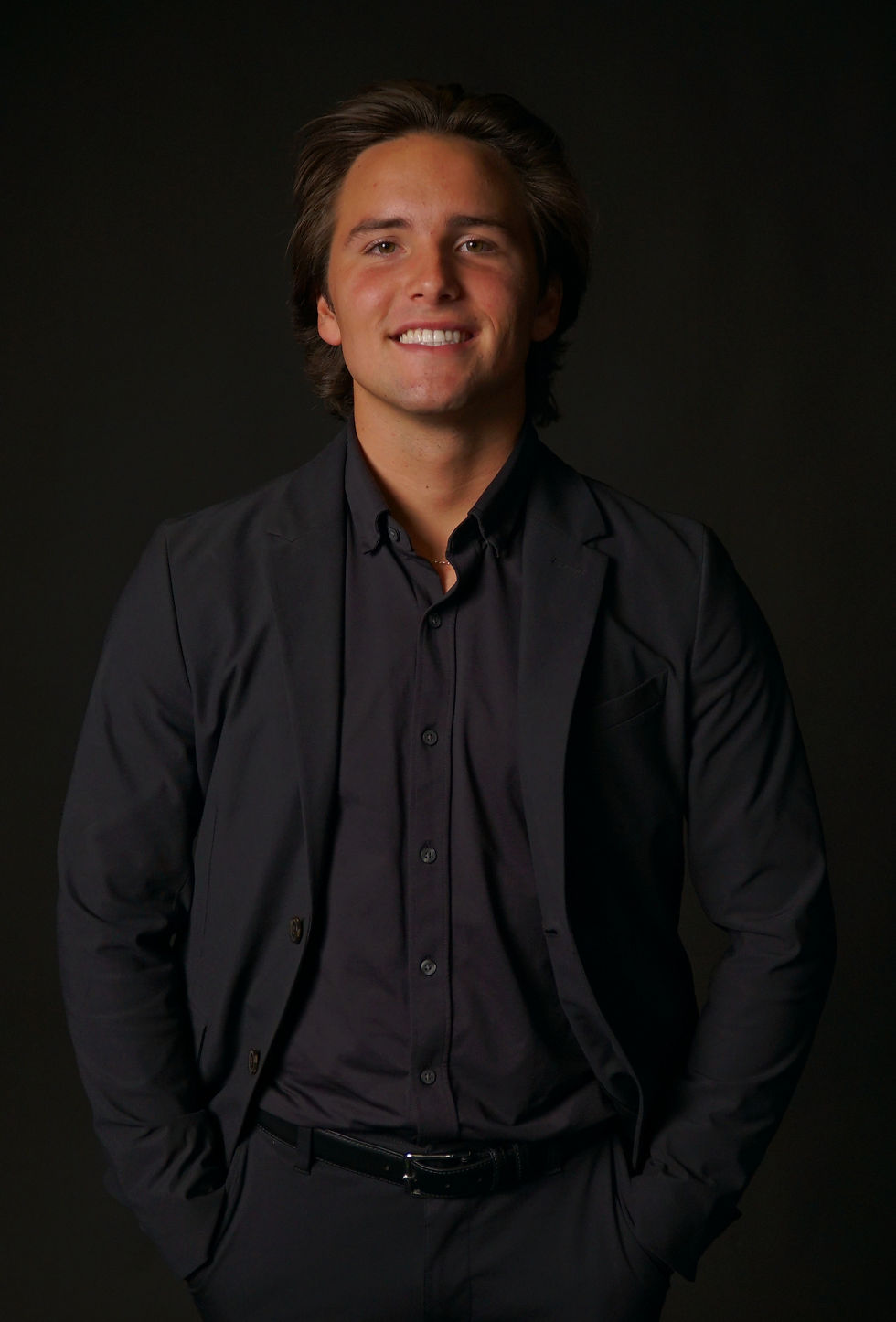 Noah DellaSala in a black suit and shirt, hands in pockets, against a dark background. The mood is confident and relaxed.
