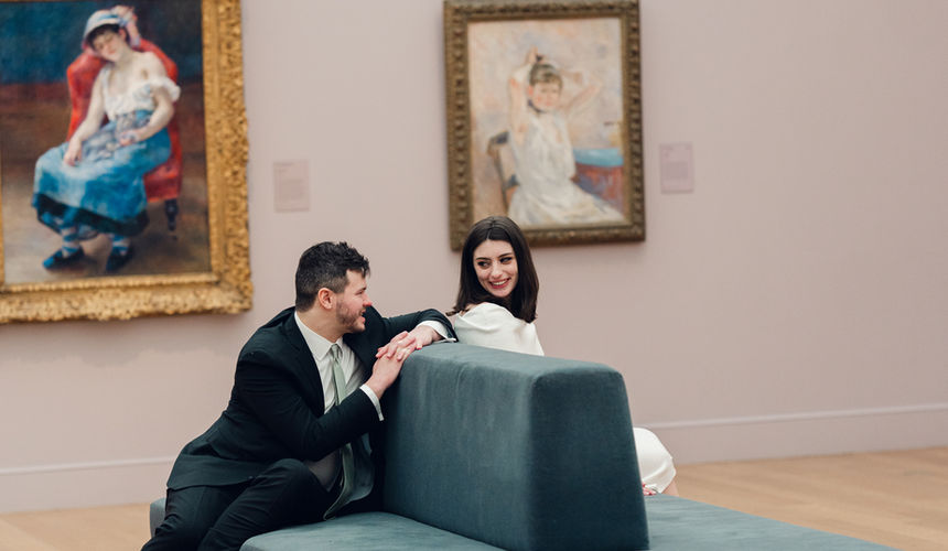 Kierstan and Kyle walking through the gallery at The Clark Art Institute, she is wearing a white knit dress with white short boots, he is in a black suit with a white collared shirt and sage green tie. They are looking at each other and smiling.