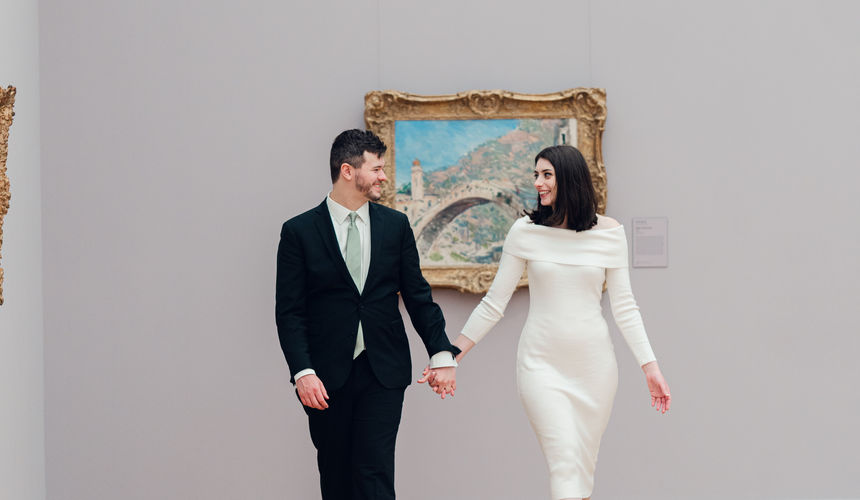 Kierstan and Kyle walking through the gallery at The Clark Art Institute, she is wearing a white knit dress with white short boots, he is in a black suit with a white collared shirt and sage green tie. They are looking at each other and smiling.