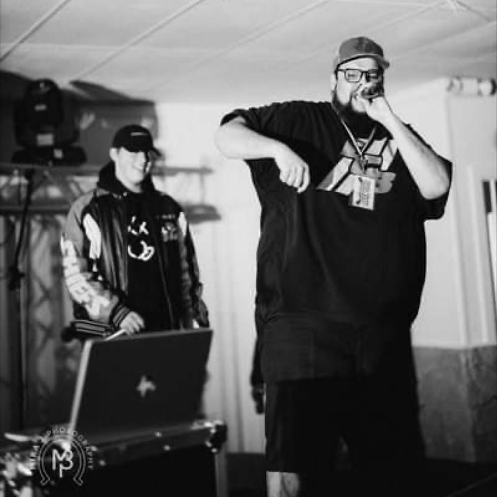 Black-and-white indoor stage with two men performing, one rapping into a mic while the other stands behind a laptop.
