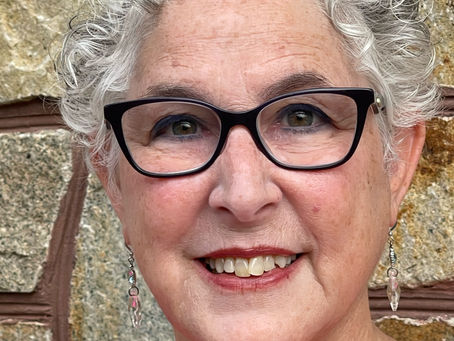 Smiling older woman with curly gray hair, black glasses, and pink top poses before a stone wall.