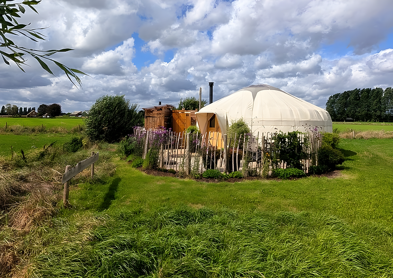 Yurt Venus in een nazomers weiland, terras met hek en bloemenrand en een Hollandse wolkenlucht erboven.