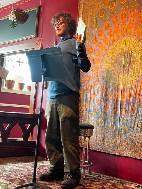 A young man stands at a reading podium holding a book while speaking to attendees during the literary tribute at Lubec Brewing Company.