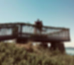 A couple stands with an officiant on a wooden lookout deck decorated with greenery, holding their wedding ceremony above the shoreline on a clear, sunny day.