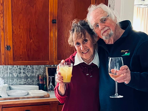 Patricia and friend standing together in a kitchen, holding drinks beside a table filled with food for a Memorial Day gathering.