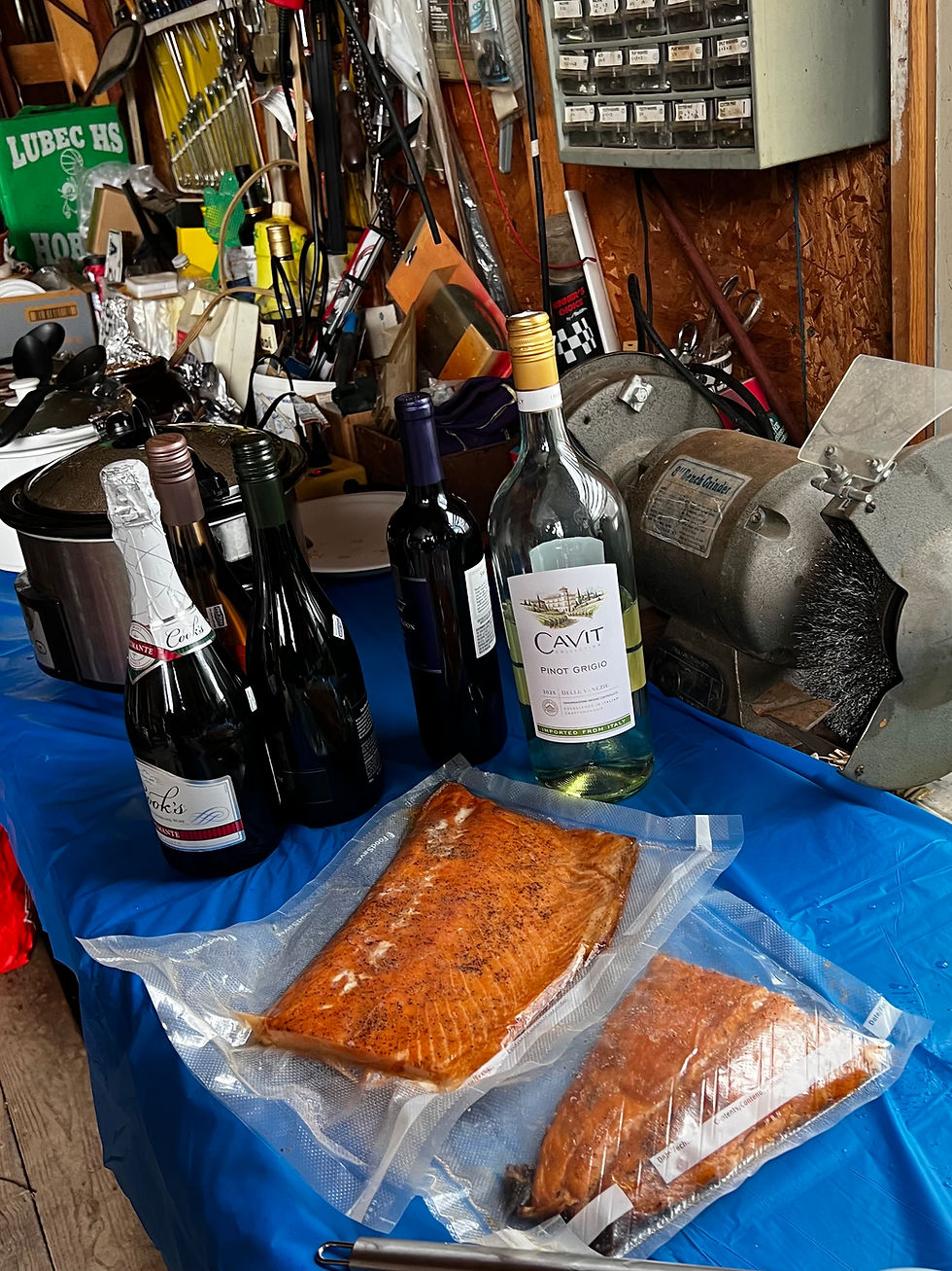 Wine bottles, sparkling beverages, and smoked fish displayed on a blue-covered table inside a garage party setting.