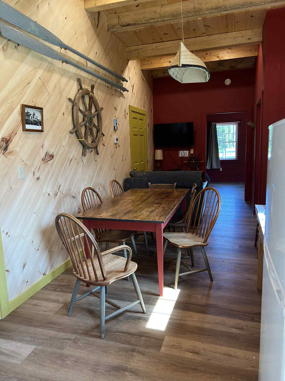 A rustic dining space with wooden table, ship’s wheel on the wall, and oars mounted above in a high-ceilinged room.