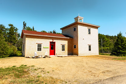 Cedar-shingled building with red door and lighthouse tower, surrounded by pine trees under a clear sky.