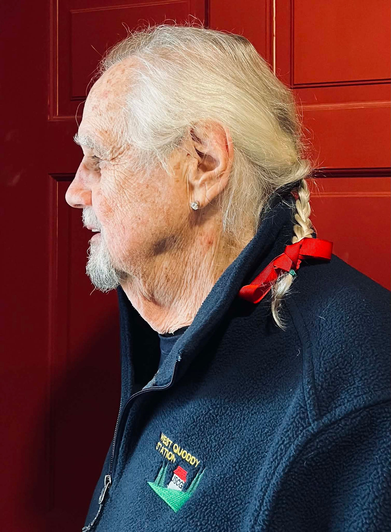 Side profile of Bill with a braided ponytail tied with a red ribbon while standing in front of a red door.