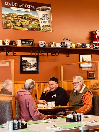 A group of older men sit at a booth in a cozy diner, drinking coffee and talking beneath shelves of mugs and local décor.
