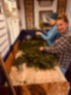 Two women smiling while preparing fir branches for wreath making inside a cozy porch setup.