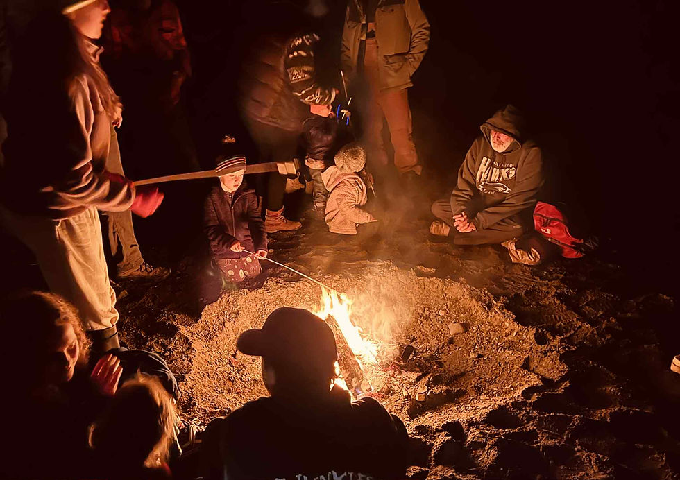 Children and adults roast marshmallows at a bonfire on the beach during Halloween festivities.