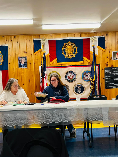 Two women seated at a table inside American Legion Post 65, organizing tickets and paperwork beneath American Legion banners and flags.
