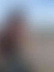 Man holding a clam rake and bucket stands on a wide sandy beach under a blue sky.