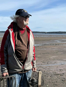 Man holding a clam rake and bucket stands on a wide sandy beach under a blue sky.