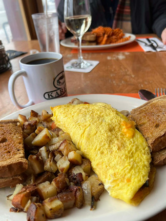 A diner breakfast plate with a cheese omelet, home fries, toast, and a coffee mug on a wooden table, with other meals in the background.