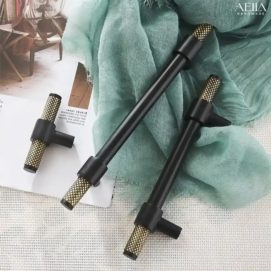 มือจับเฟอร์นิเจอร์สีดำหลายแบบวางบนผ้าสีเขียวและมีป้าย AELLA HARDWARE L3070