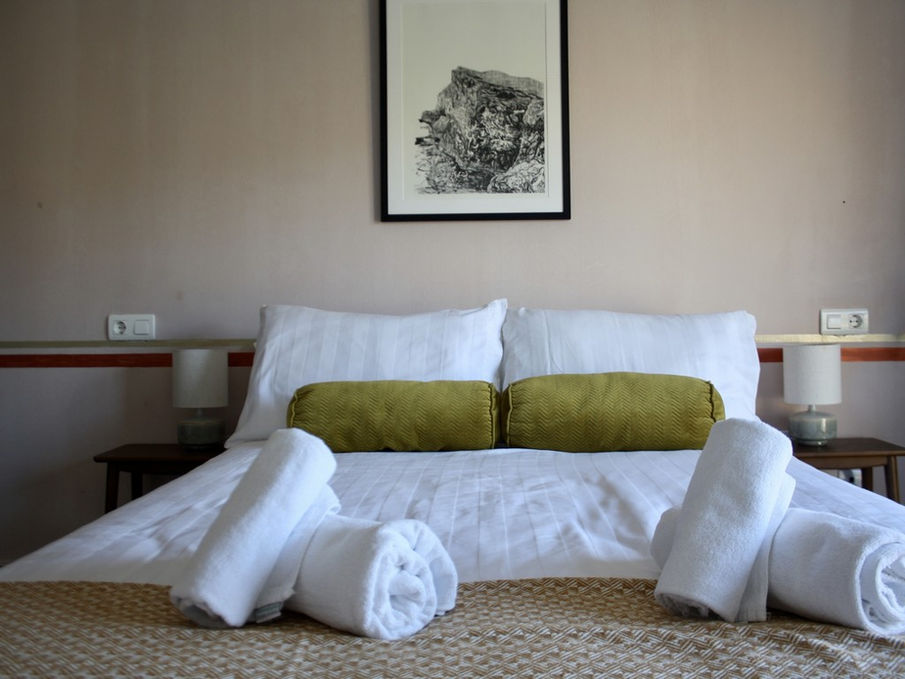 Habitación con cama hecha, sábanas blancas, dos almohadas color oliva y toallas enrolladas.