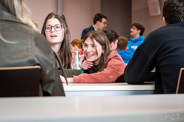 Two students sitting behind a desk look over toward a third students backing from the camera