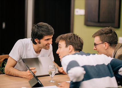 Three students talk to each other over a wooden desk and smile confidentially