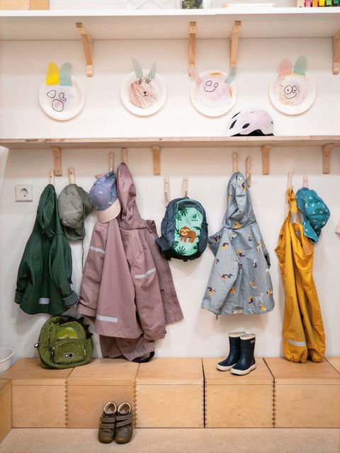 Garderobenbereich in einer Kita mit Kindermänteln, Rucksäcken und Gummistiefeln an Haken und Sitzboxen aus Holz. Darüber hängen Kinderzeichnungen auf Papptellern an der Wand.