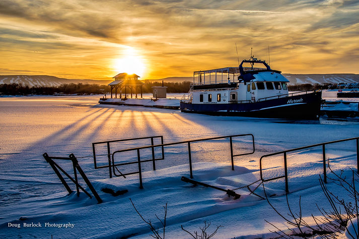 Winter scene of Collingwood Chartered boat and snow covering the water overlooking the yellow sky sunset and ski hills in background
