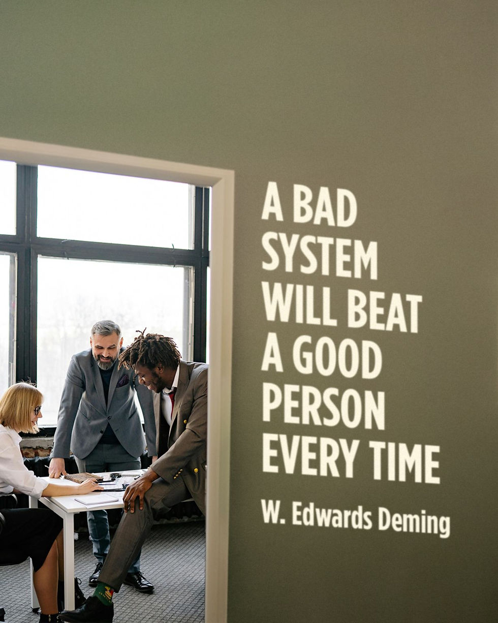 Team meeting in a bright office beside wall quote: A BAD SYSTEM WILL BEAT A GOOD PERSON EVERY TIME, W. Edwards Deming