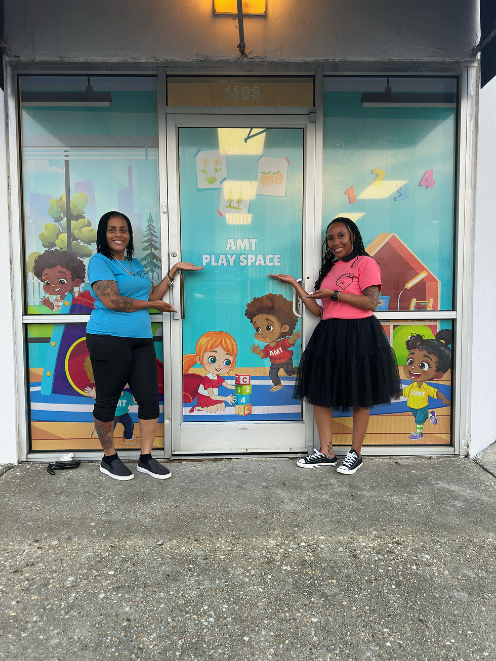 Two women smile, pointing at a door labeled "AMT Play Space" with colorful cartoon kids. They're outside a building with a playful vibe.