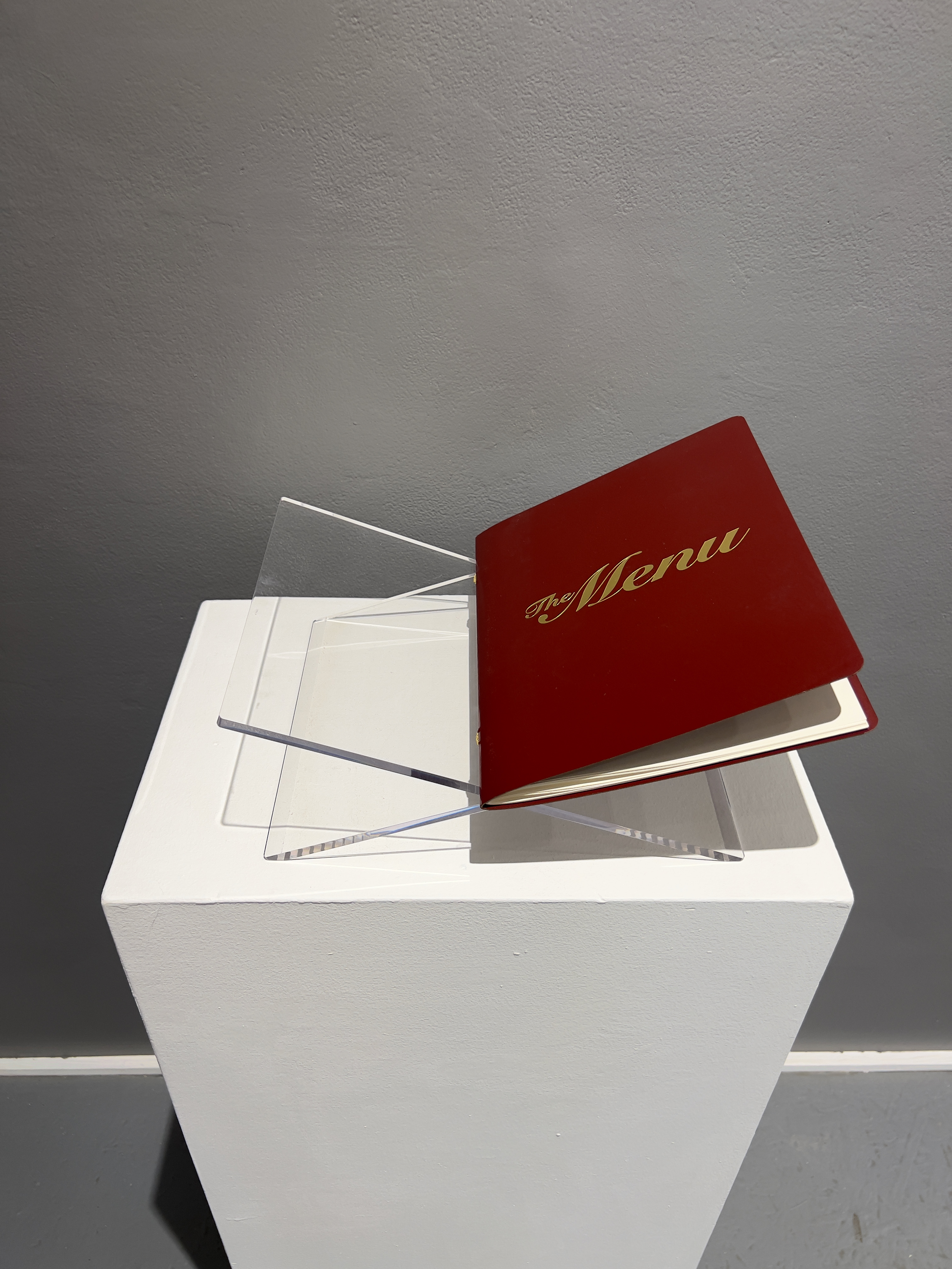 Pedestal display view of the artist's book, The Menu. The Menu is printed in gold foil lettering and the cover is burgundy.  Title is The Menu.  