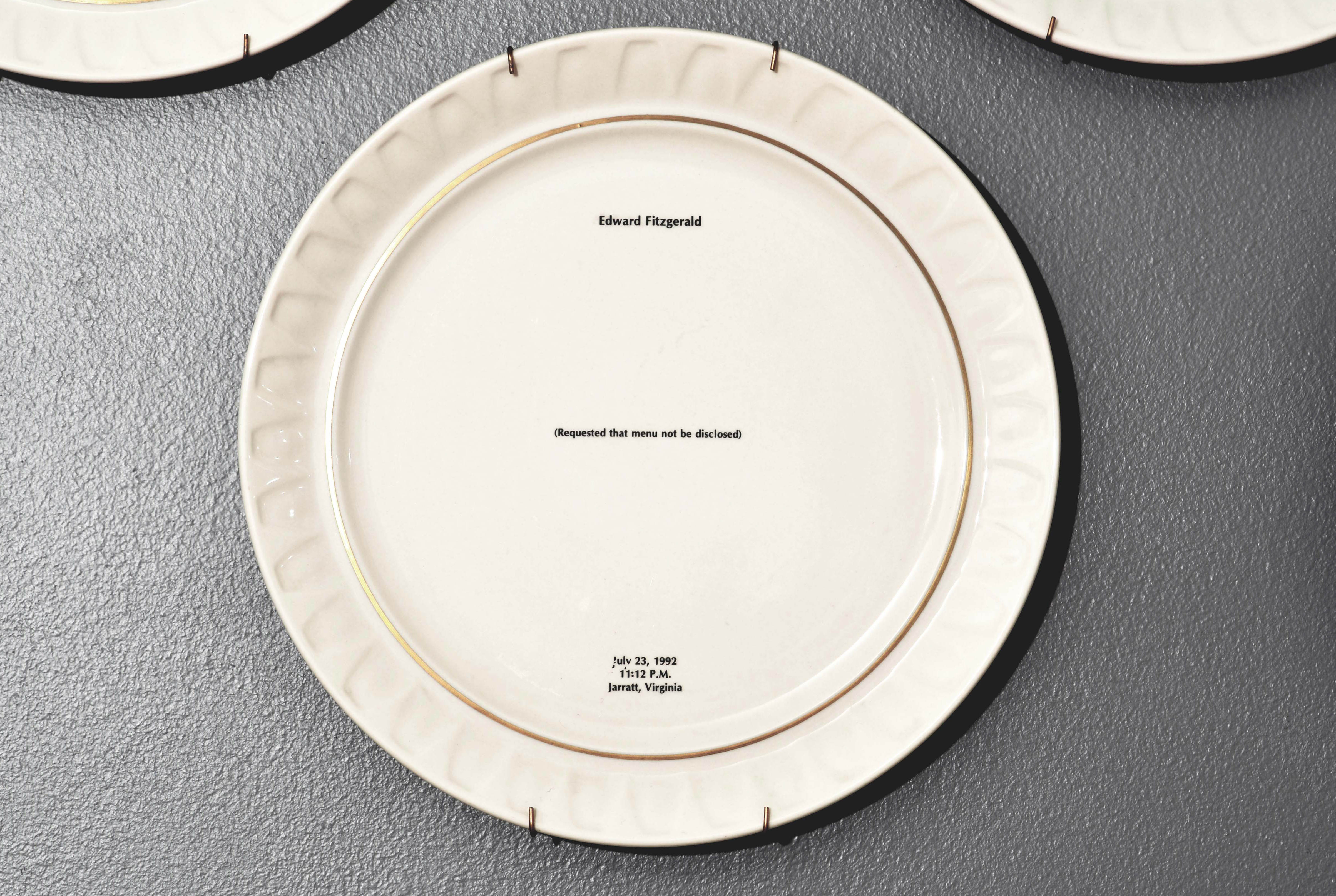 Bone china dinner plate, with gold trim and text printed in center. Text includes a description of the last meal requested and served to someone prior to their execution. The name of the person, time, date and place of execution is also printed on the plate. Detail from installation.