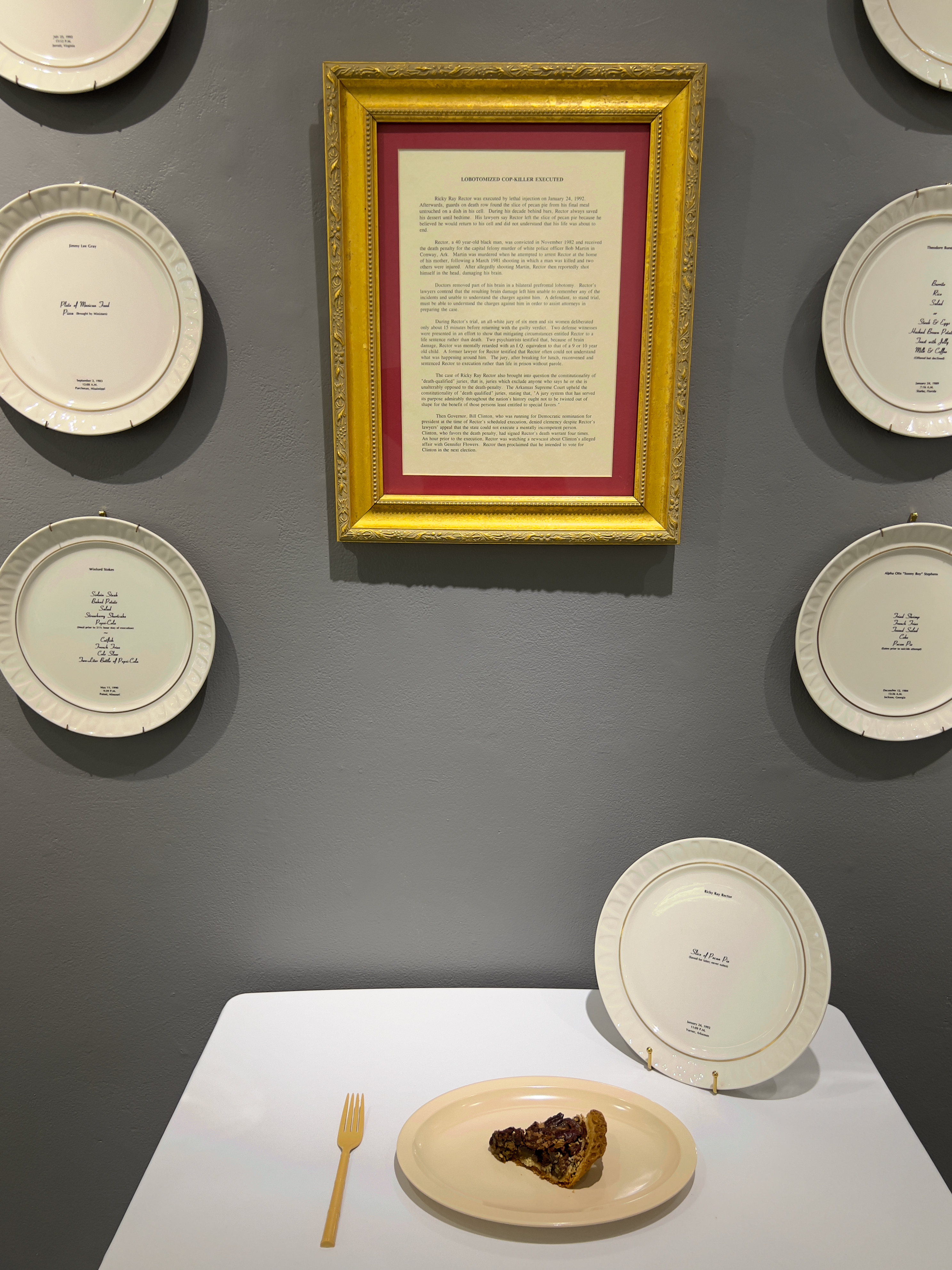 Mixed media installation which includes small table covered with white tablecloth.  One slice of pecan pie sits on a plastic dish.  A bone china dinner plate is standing. 18 bone china dinner plates hang on a gray wall.  Gold framed story about Ricky Ray Rector hangs centered on the wall behind the table.