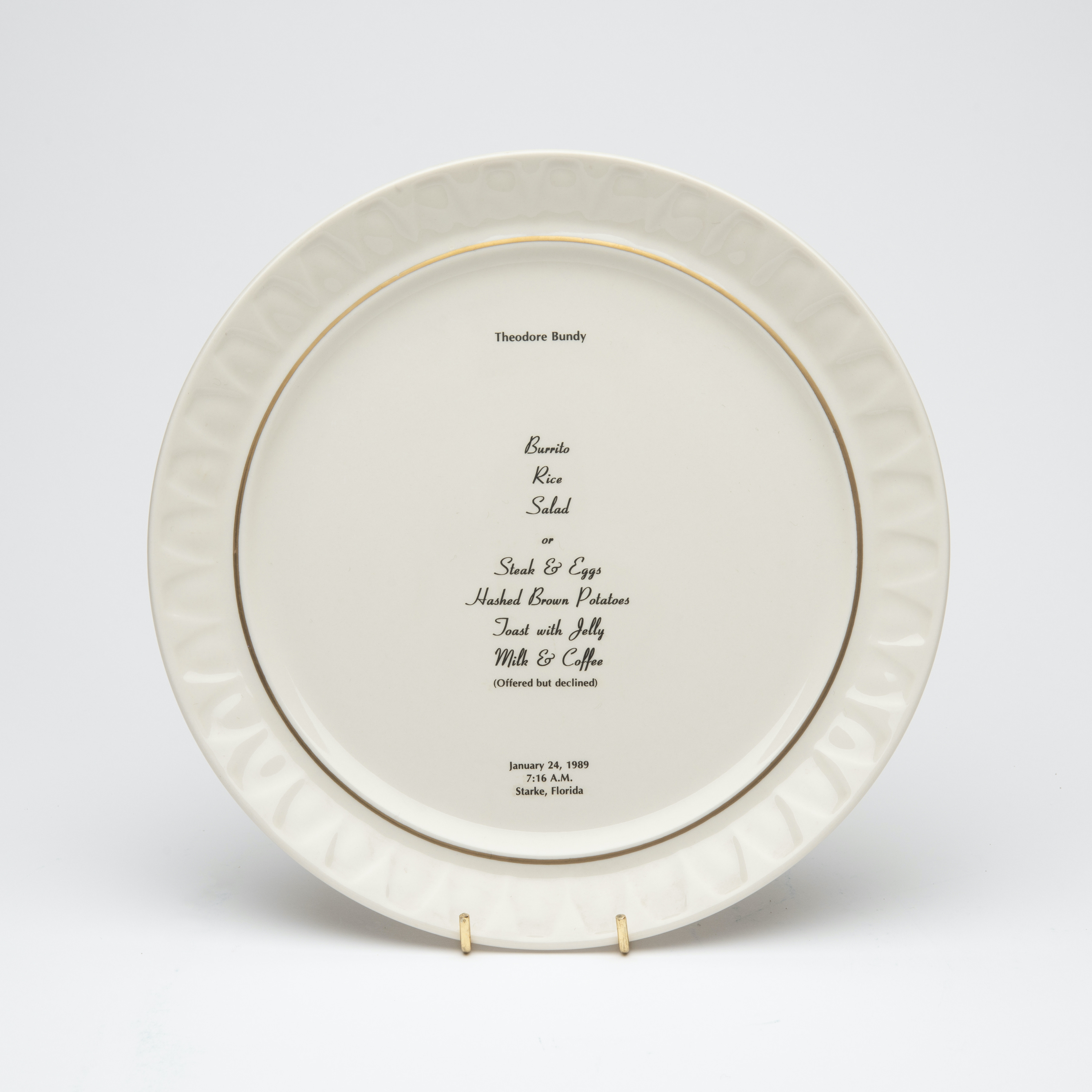 Bone china dinner plate, with gold trim and text printed in center. Text includes a description of the last meal requested and served to someone prior to their execution. The name of the person, time, date and place of execution is also printed on the plate.