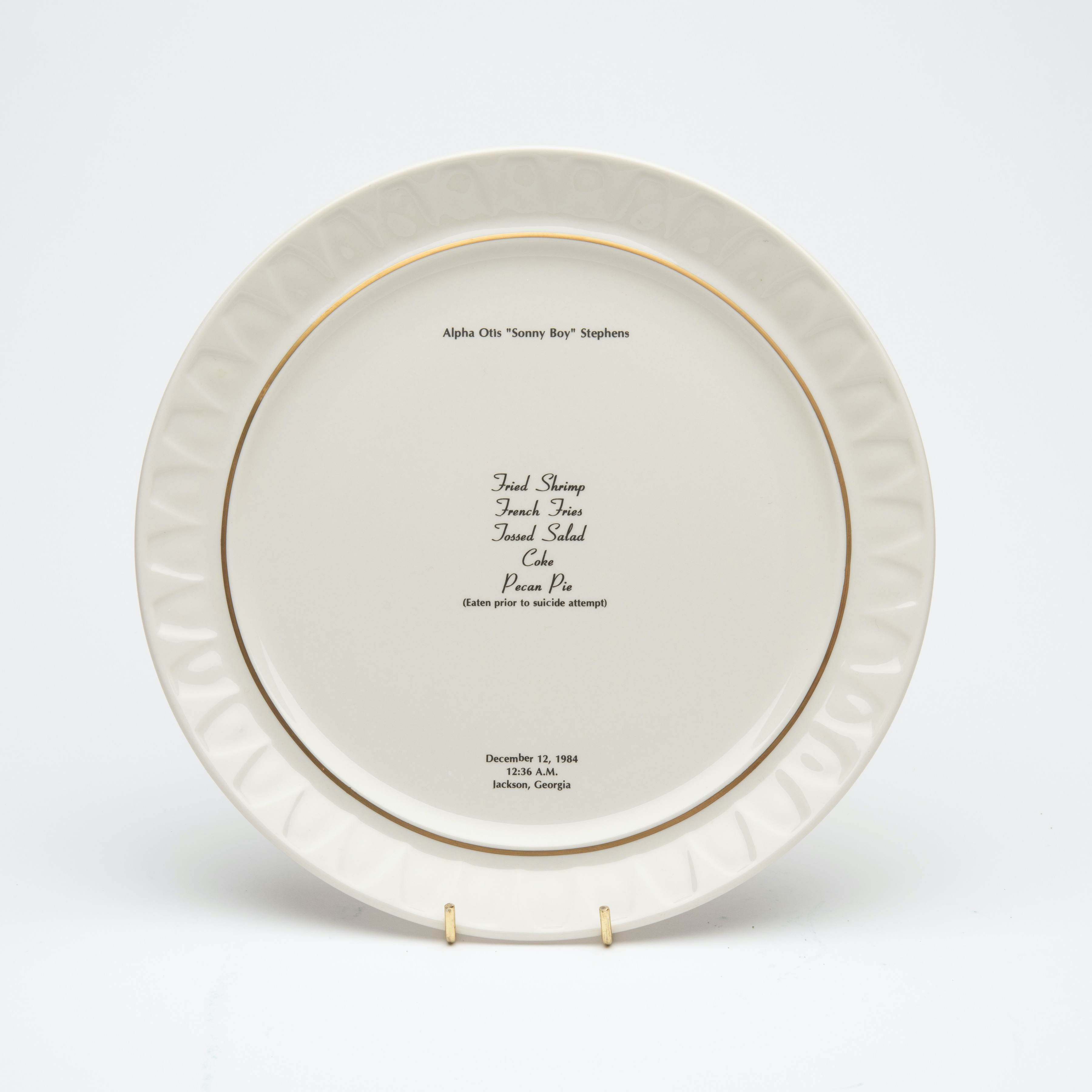 Bone china dinner plate, with gold trim and text printed in center. Text includes a description of the last meal requested and served to someone prior to their execution. The name of the person, time, date and place of execution is also printed on the plate.