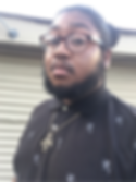 Justin, a Black person, looks at the camera. He wears a black button-up shirt with white lion pattern, a Prince symbol necklace and brown glasses. His hair is pulled back and he has a beard and mustache.