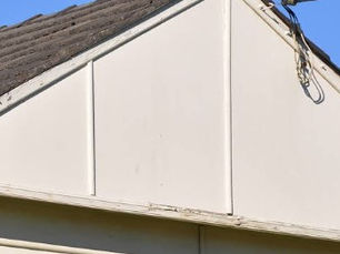 A close-up photograph of an older Australian house showing a faded white/cream asbestos-cement (AC) fibro gable end panel, separated by vertical timber joiner batten strips.