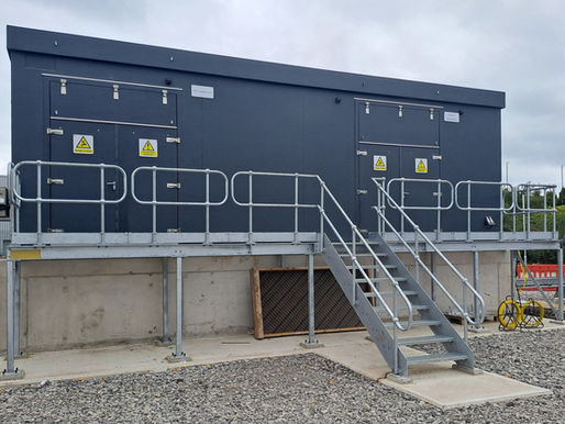 Gray GRP substation by Hill Grwp with stairs and handrails on a gravel surface. "Danger of Death" signs on the doors. Overcast sky.