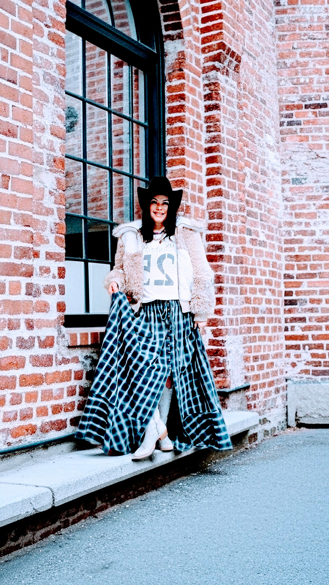 Woman wearing a western boho outfit with a faux fur jacket, graphic tee, flowing tie-dye skirt, wide-brim cowboy hat, and boots, standing against a brick building