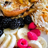 A white oval plate featuring two thick slices of golden-brown brioche French toast, topped with melted butter and syrup. The plate is beautifully styled with a heap of fresh raspberries, blackberries, and sliced bananas. On the side are crispy shredded hash browns and a strip of turkey bacon, set against a neutral, rustic background.