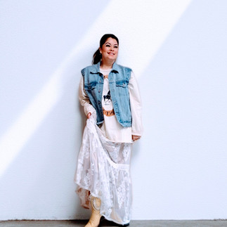 A Boho-Western layered look with a denim vest over a white lace maxi dress and a graphic tee, completed with yellow-colored cowboy boots.