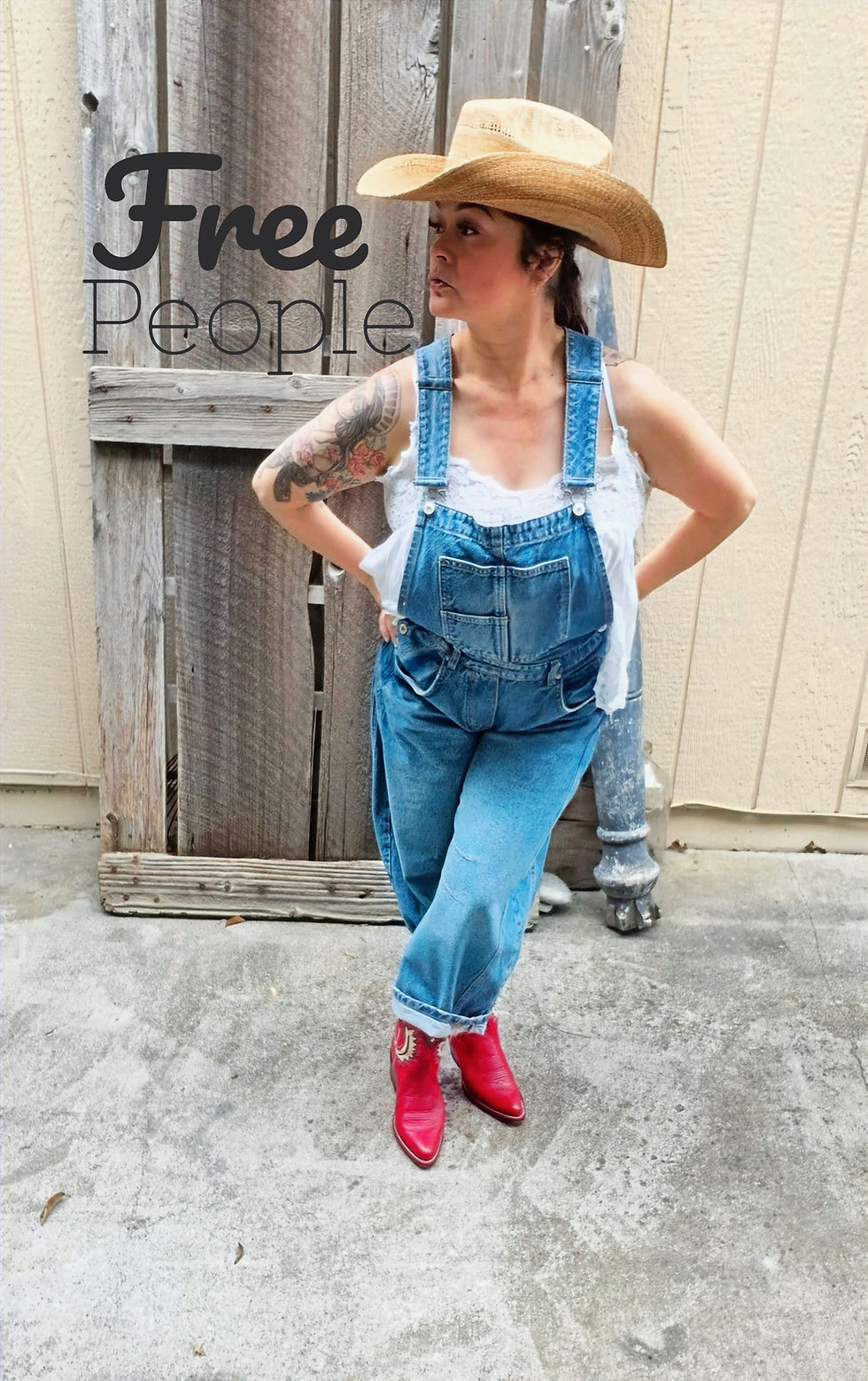 Woman in denim overalls poses confidently on a patio. Wooden fence with a wreath and string lights in the background. Overcast mood.
