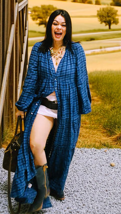 A close-up shot of a woman wearing a blue and white vertical striped dress with a ruffled neckline and tie-front detail. She is leaning forward, highlighting the breezy fabric and the subtle appearance of dark bloomers through the side slits of the garment against a white wood background.