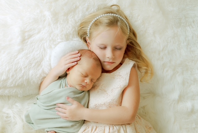 Sleeping newborn cradled in their sister’s arms during a tender moment captured by Suzi Brown Photography